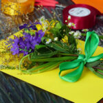 9. Na zdjęciu widzimy bukiet z kwiatów na żółtej kartce. W tle leżą czerwone i żółte wstążki w szpulce.