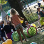 2. Na zdjęciu widzimy grupę dzieci wraz z rodzicami. Dzieci bawią się na trawie. W tle stoi wigwam i tor przeszkód. Na trawie leżą balony w kolorze żółtym.
