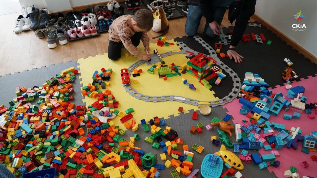 Alt=”Strefa Lego Masters w CKiA. Na kolorowych matach rozrzucone setki kolorowych klocków LEGO. Chłopiec z tatą składają tory kolejowe. Za nimi ustawione pod ścianą zdjęte kilkadziesiąt par butów.”