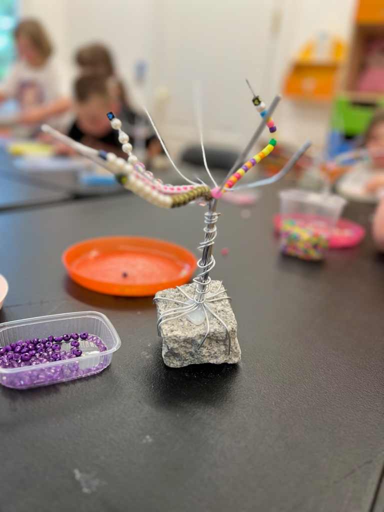 Alt=”Warsztaty kreatywne dla dzieci. Na zdjęciu widzimy wykonywane przez uczestnika drzewko. Podstawą jest kamień, od którego odchodzą druciki imitujące gałęzie. Dzieci dekorują gałęzie kolorowymi ozdobami.”